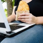 Girl-eats-pie-and-looks-at-laptop-6-types-of-obesity-school-nutrition-academy-Wellness-Consulting
